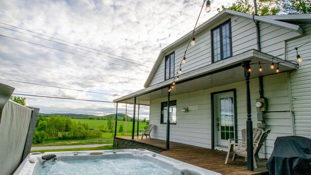 A house with a hot tub on the deck, a covered porch, and a view of a green landscape. - Les Chalets Tourisma - La Campagnarde (Saint-Leonard-de-Portneuf)