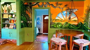 A room with a wooden floor, a round table with two chairs, a green bookshelf, and a mural of a sunset. - Creekside Paradise • Sunsets, Pool, Spa, Dock, Kayaks (Tybee Island)