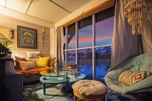 A cozy living room with a large window, a wicker chair, a sofa with cushions, a glass coffee table, and a framed picture on the wall. - Creekside Paradise • Sunsets, Pool, Spa, Dock, Kayaks (Tybee Island)