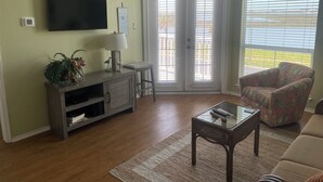 En stue med et fladskærms-tv, en lampe, en dekorativ vægdekoration, en bænk, et glas-sofabord og en mønstret lænestol.
