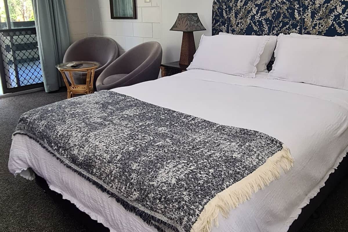 Une chambre d’hôtel avec un grand lit, un ventilateur de plafond, une petite table avec une chaise, une lampe et une vue sur l’extérieur par une fenêtre avec des rideaux.