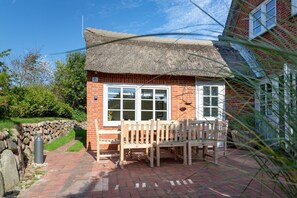 A brick building with a thatched roof, a stone wall, and a paved patio with wooden furniture.