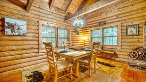 A log cabin interior with a wooden dining table, four chairs, two windows with blinds, a chandelier, and rustic decor including a wagon wheel and framed pictures.
