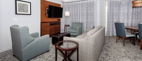 Una sala moderna con un sofá, sillones, una mesa de centro de cristal, un televisor de pantalla plana, una lámpara y un comedor con sillas y una mesa.