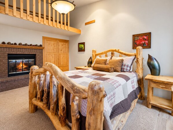 A bedroom with a wooden bed frame, a fireplace, and a high ceiling with a wooden railing.