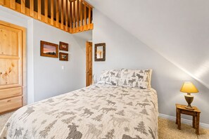 A bedroom with a sloped ceiling, a bed with a moose print bedspread, a wooden wardrobe, and a small bedside table with a lamp.