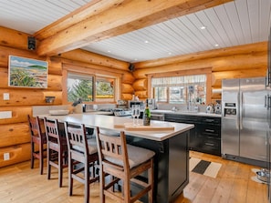 Une cuisine rustique aménagée dans une cabane en rondins, avec des murs en bois, un îlot central doté de tabourets de bar, un réfrigérateur en acier inoxydable et une fenêtre donnant sur un paysage verdoyant.