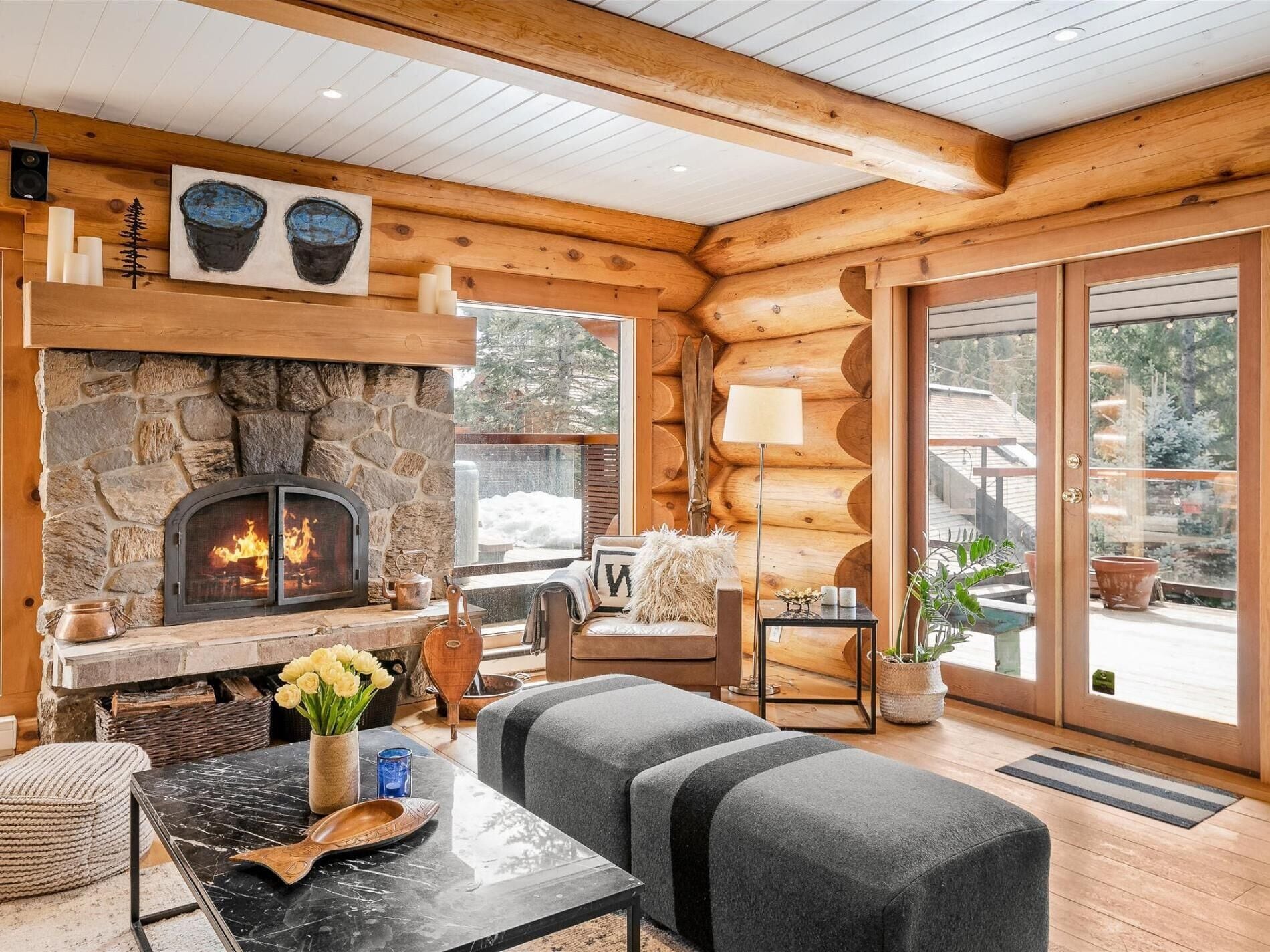 L’intérieur chaleureux d’un chalet en bois, avec une cheminée en pierre, un coin salon confortable et de grandes fenêtres donnant sur le paysage extérieur.
