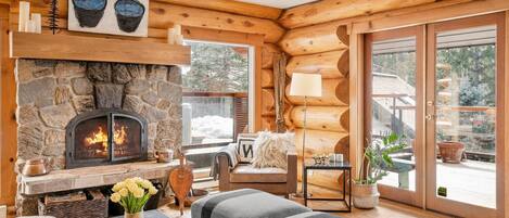 L’intérieur chaleureux d’un chalet en bois, avec une cheminée en pierre, un coin salon confortable et de grandes fenêtres donnant sur le paysage extérieur.