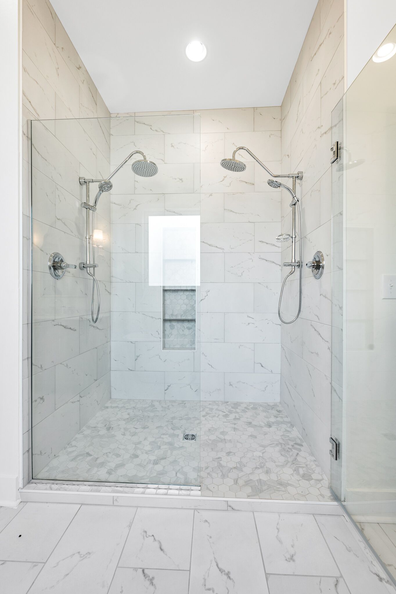 Une douche fermée par des parois de verre, avec un sol et des murs carrelés, une petite fenêtre et un luminaire au plafond.