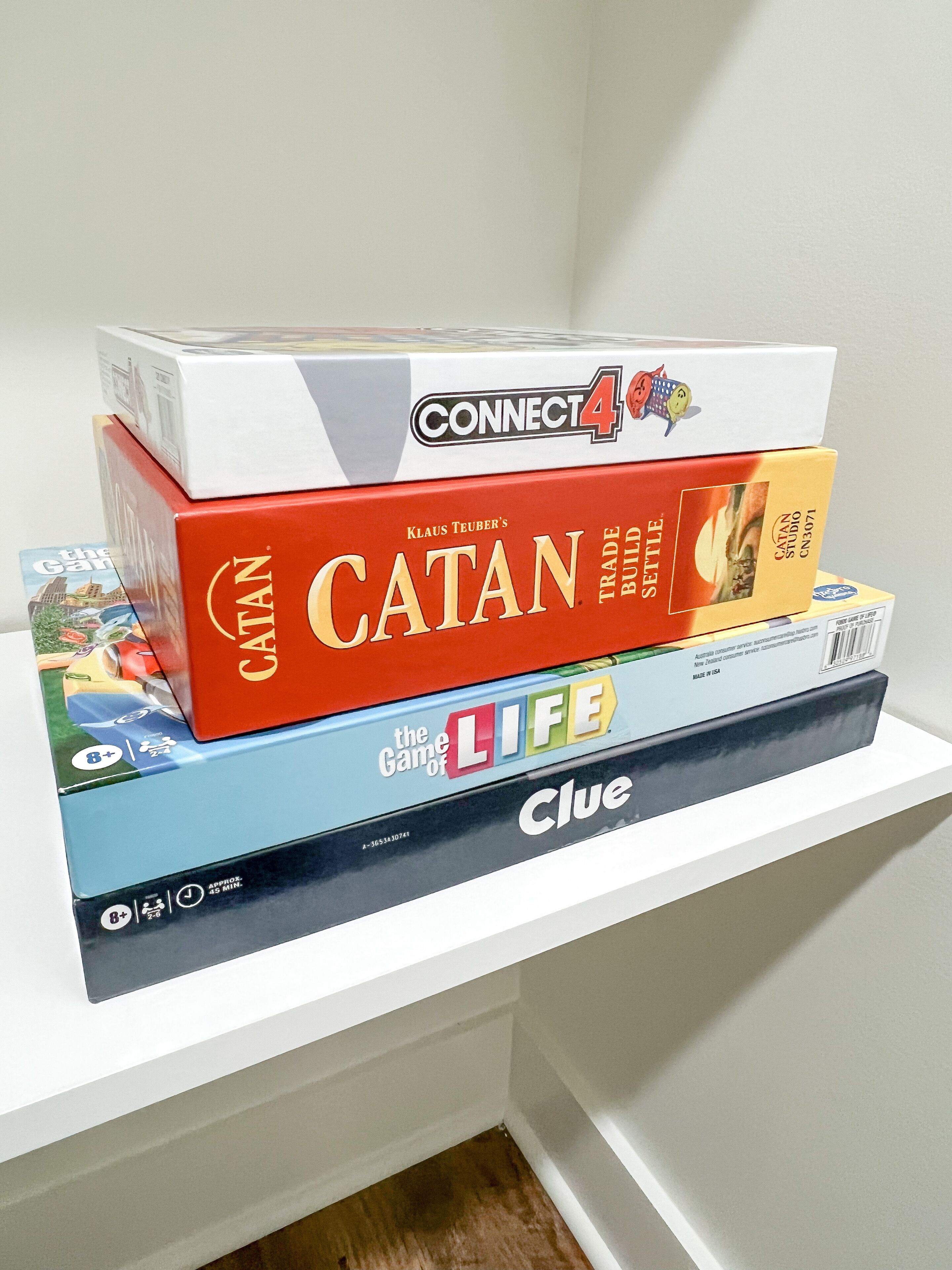 Une étagère avec quatre jeux de société : Connect 4, Catan, La Vie est un jeu et Cluedo.