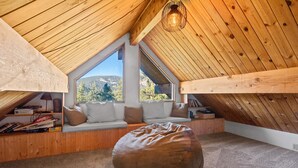A cozy attic room with wooden beams, a large bean bag, and a view of a forested hillside. - TreeHouse on Rainbow - Views, Fireplace, Deck, Barbecue (Big Bear City)