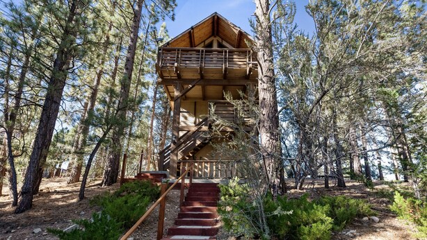 A wooden cabin with a staircase leading to a balcony, surrounded by trees. - TreeHouse on Rainbow - Views, Fireplace, Deck, Barbecue (Big Bear City)
