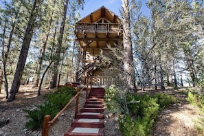A wooden cabin with a staircase leading to a balcony, surrounded by trees. - TreeHouse on Rainbow - Views, Fireplace, Deck, Barbecue (Big Bear City)
