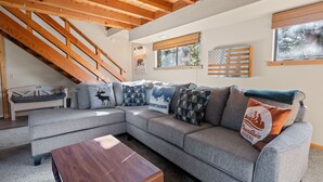 A modern living room with a sectional sofa, wooden beams, a staircase, and a window with blinds. - TreeHouse on Rainbow - Views, Fireplace, Deck, Barbecue (Big Bear City)