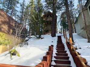 Eine mit Schnee bedeckte Holztreppe führt zu einer Hütte, umgeben von hoch aufragenden Kiefern.