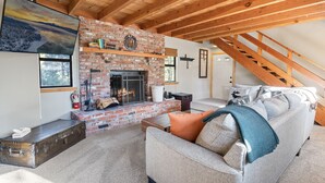 A cozy living room with a brick fireplace, a sofa with cushions, a wooden ceiling, and a staircase. - TreeHouse on Rainbow - Views, Fireplace, Deck, Barbecue (Big Bear City)