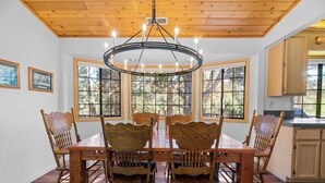 A dining area with a wooden table and chairs, large windows, and a chandelier.