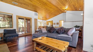 A spacious living room with a wooden ceiling, a grey sofa set, a wooden coffee table, and large windows offering views of the outdoors.