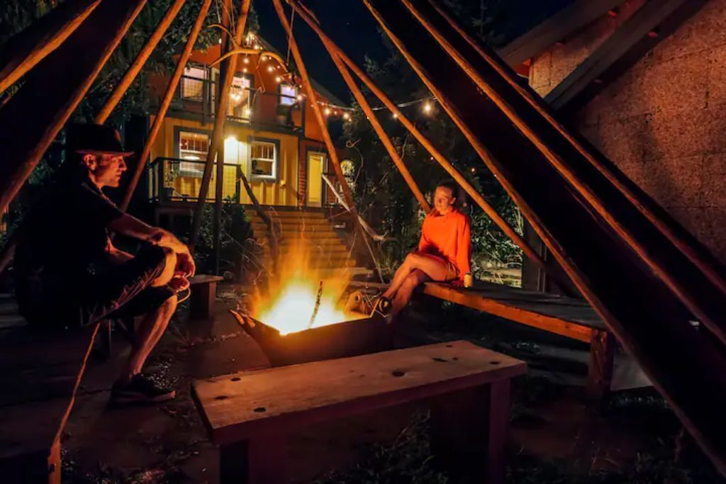Two people sitting by a campfire in an outdoor setting with a house in the background.