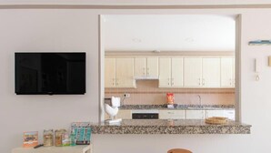Een keuken met lichte kastjes, een granieten aanrechtblad, een zwarte televisie aan de muur en een deur rechts.
