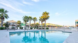 A swimming pool with lounge chairs and palm trees, surrounded by buildings with balconies and a white fence. - 2 Homes- Perfect location for large families or groups. Pool, Walk to BEACH (Port Aransas)