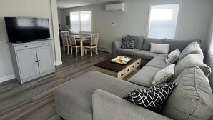 Una sala de estar moderna con un sofá gris, un televisor de pantalla plana, un comedor con mesa y sillas, y una ventana que proporciona luz natural.