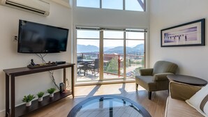 A modern living room with a large TV, a console table with plants, a round glass coffee table, a sofa, and a chair. - Stunning Lake View Condo (West Kelowna)