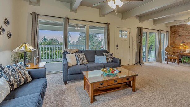 A spacious living room with a dark gray sofa, a glass coffee table, and a ceiling fan. Large windows provide ample natural light and a view of the outdoors. - Redondo Beach Bungalow (Des Moines)