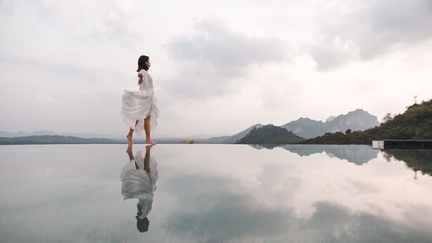 Une femme vêtue d’une robe blanche, debout sur une surface réfléchissante, avec des montagnes en arrière-plan.