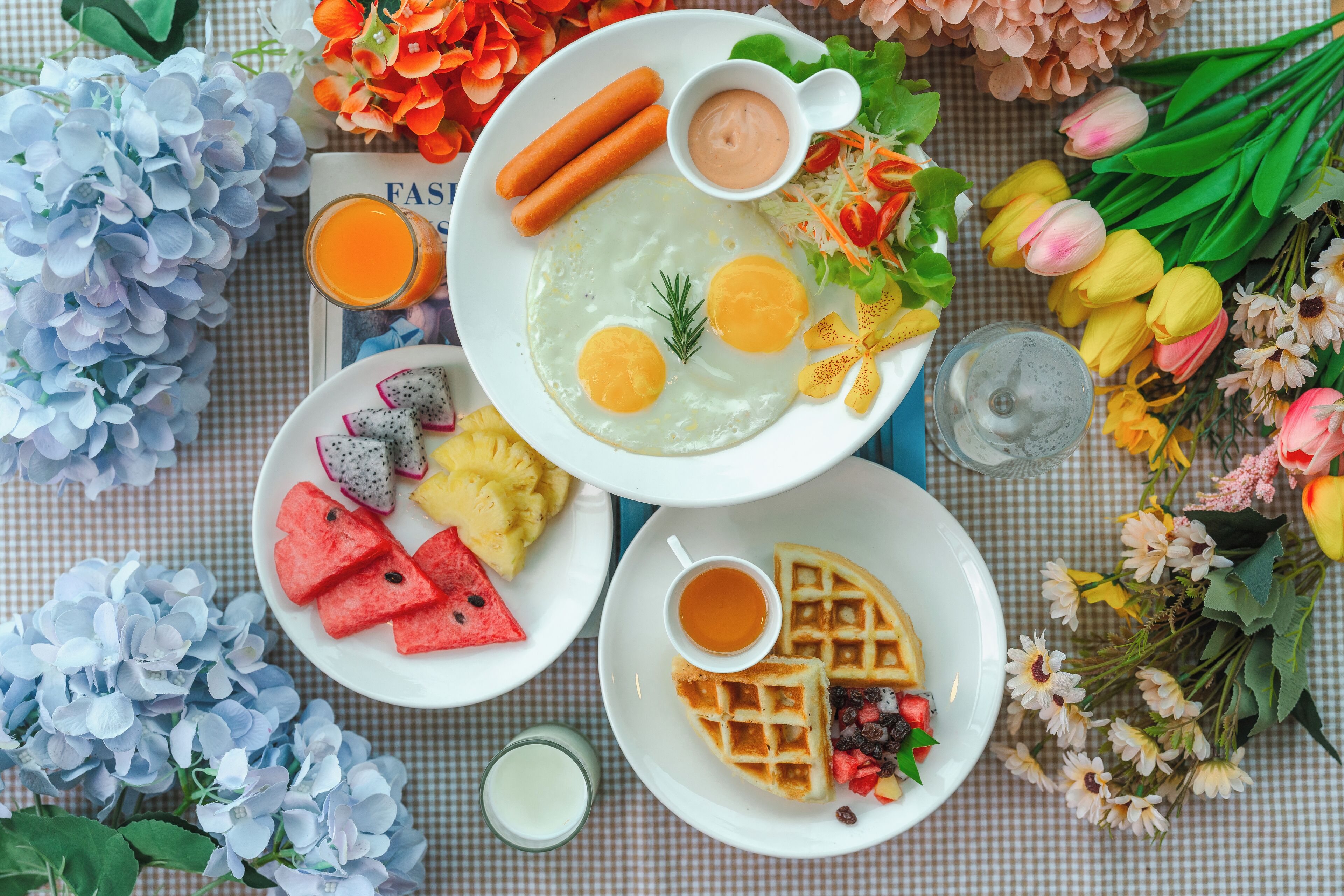 A breakfast table with various dishes including eggs, sausages, and waffles, surrounded by flowers and fruits.