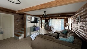 A cozy living room with a wooden ceiling, a television, a dining area with chairs, and a kitchen in the background.
