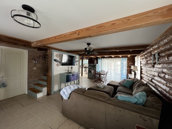 A cozy living room with a wooden ceiling, a television, a dining area with chairs, and a kitchen in the background.