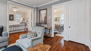 Amplia sala de estar con piso de madera, un sillón azul claro, un sofá gris oscuro y una mesa de centro de madera. Hay una puerta blanca que da a otra habitación.