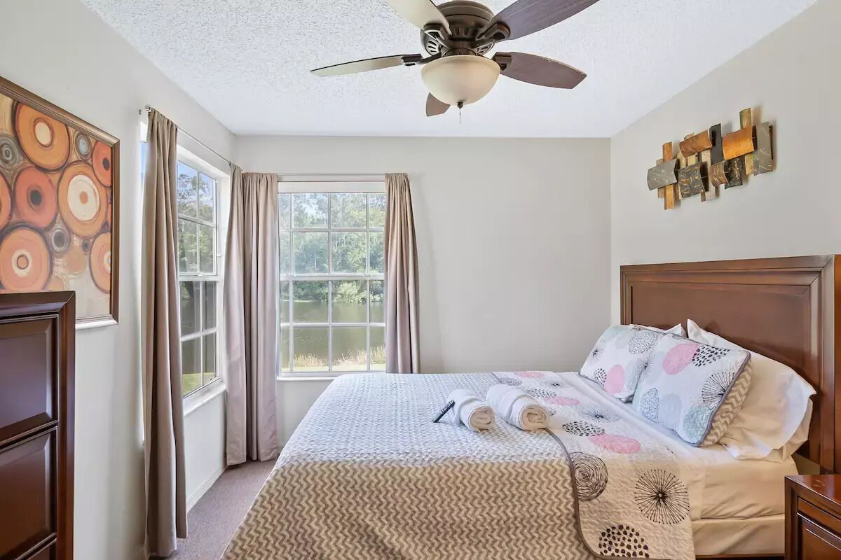 A bedroom with a bed, a ceiling fan, a window with curtains, and a wall with a decorative piece.