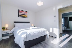 A modern bedroom with a large bed, a nightstand with a lamp, a framed picture on the wall, and a view into another room. - Stunning 2-Bed House in Manchester with Canal view (Salford)