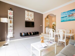 Un salon moderne avec un téléviseur à écran plat, une bibliothèque, une table basse avec des livres et un tableau représentant un paysage côtier.