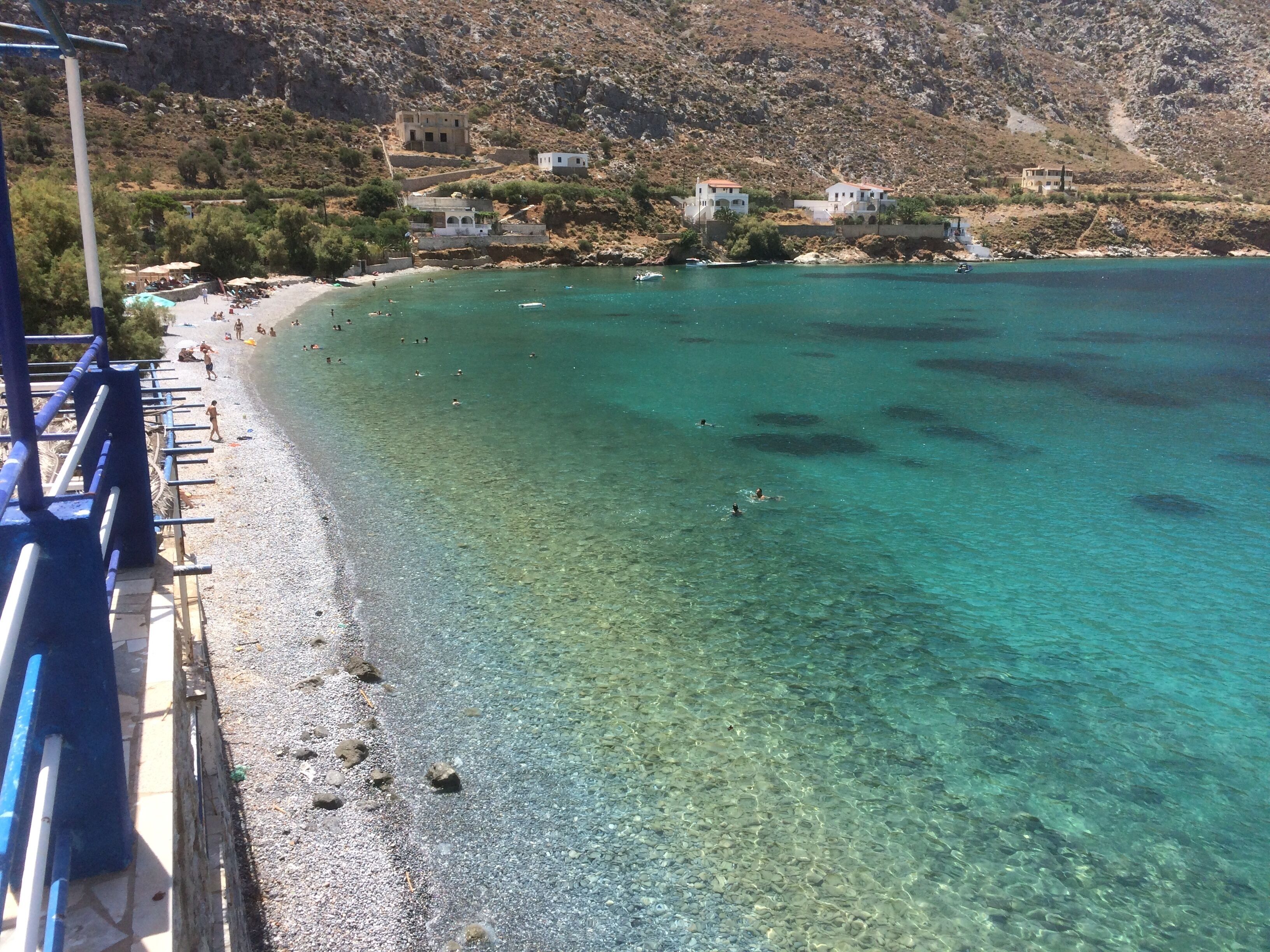Une plage côtière aux eaux turquoise et limpides, avec un terrain rocheux et quelques bâtiments dispersés.