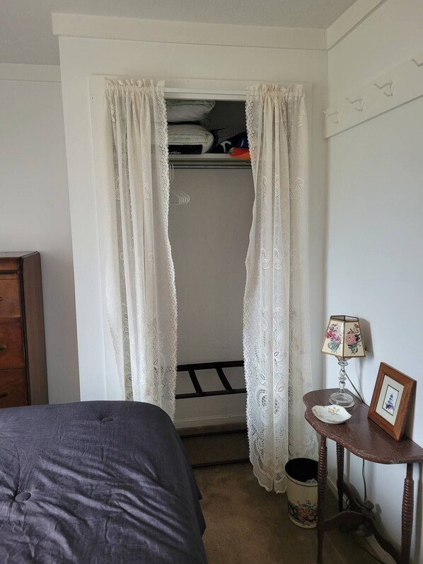 A bedroom with a bed, a small table with a lamp, a wooden dresser, and a closet with hanging clothes.
