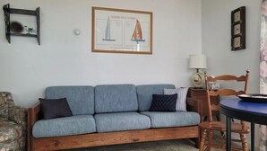 Un salon avec un canapé bleu, une chaise en bois, une petite table et un tableau encadré représentant des voiliers accroché au mur.