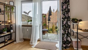 Una sala de estar moderna con piso de madera, un alfombra blanco y esponjoso, una estantería de metal negro con plantas, una puerta de vidrio que da a un balcón con vista a árboles y una valla de madera.