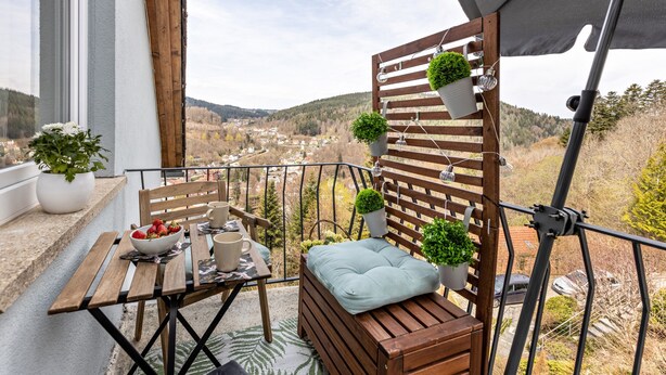 Un balcón con mesa de madera, sillas, un banco acolchado, plantas en macetas y vistas a un pueblo y colinas.