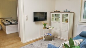 A compact living space with a bed, a TV mounted on the wall, a cabinet with glass doors, and a small table with a vase of flowers. - Apartment "Quartier 3" with Wi-Fi (Zeven-Brauel)
