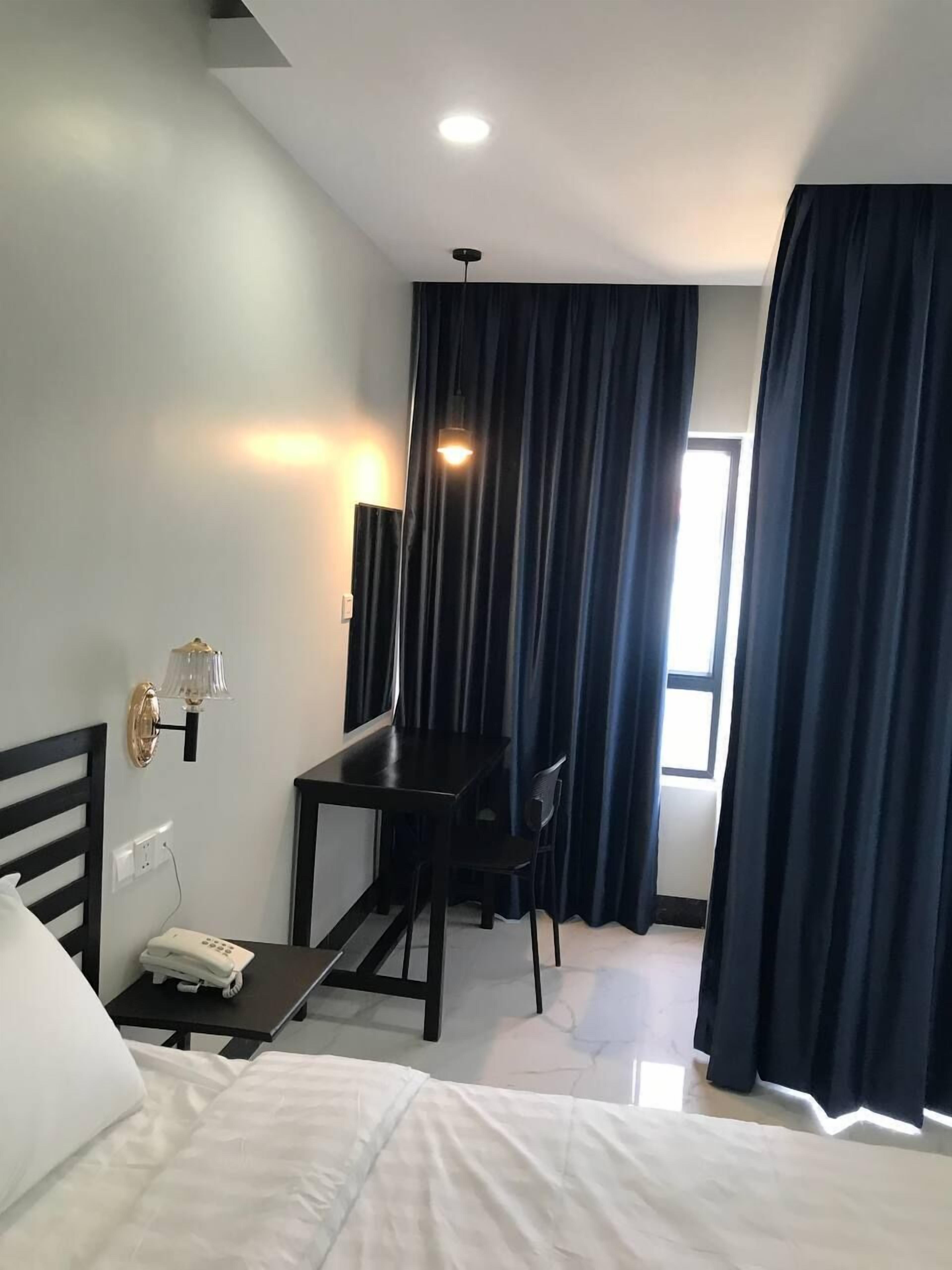 A hotel room with a bed, a desk with a chair, a lamp, a telephone, and a window with curtains.