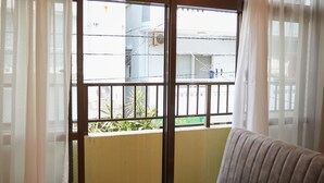 Una habitación con una puerta corrediza de vidrio que da a un balcón. El balcón tiene barandilla de metal y ofrece vistas de edificios y un ventilador en el exterior.