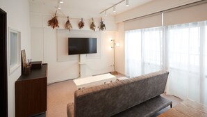 Una sala de estar moderna con un sofá gris, un televisor de pantalla plana, un escritorio de madera y un ventanal con cortinas translúcidas.