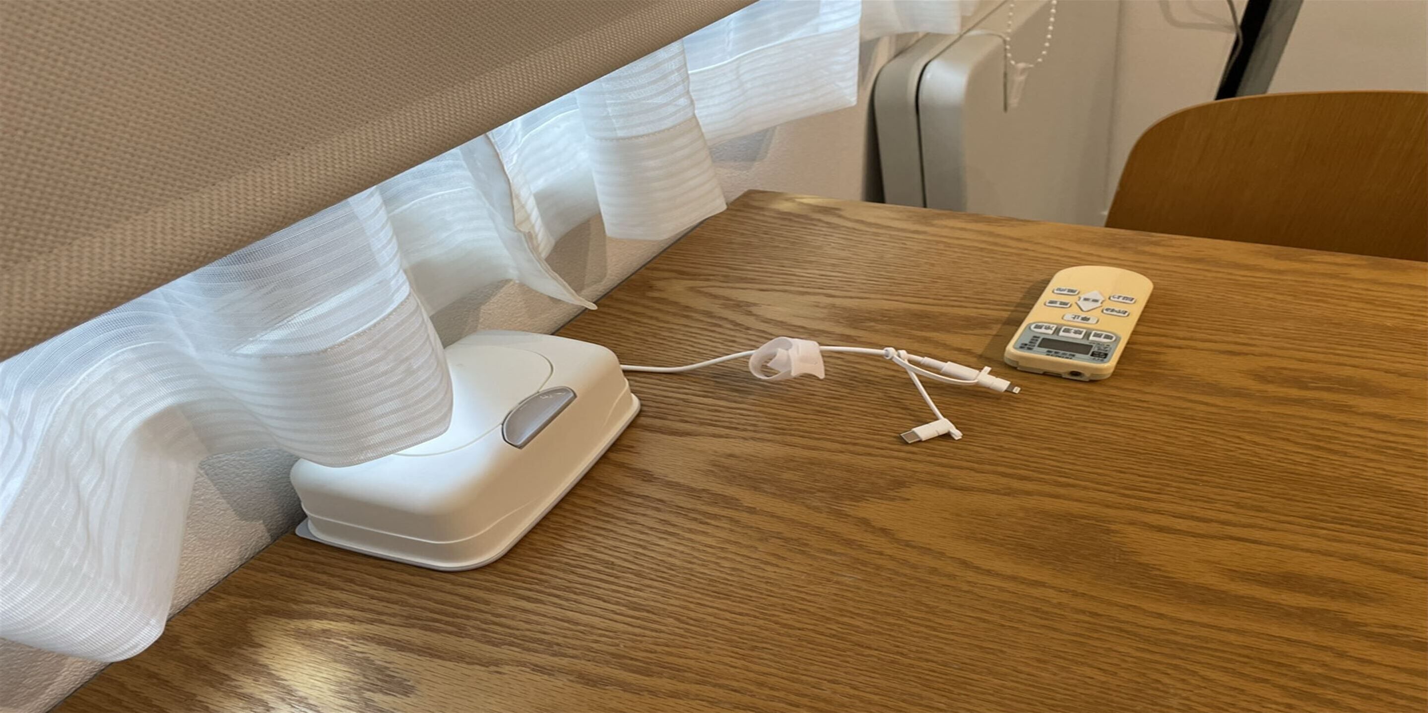 A bed with white sheets and a white bedside table with a white device and charging cables.