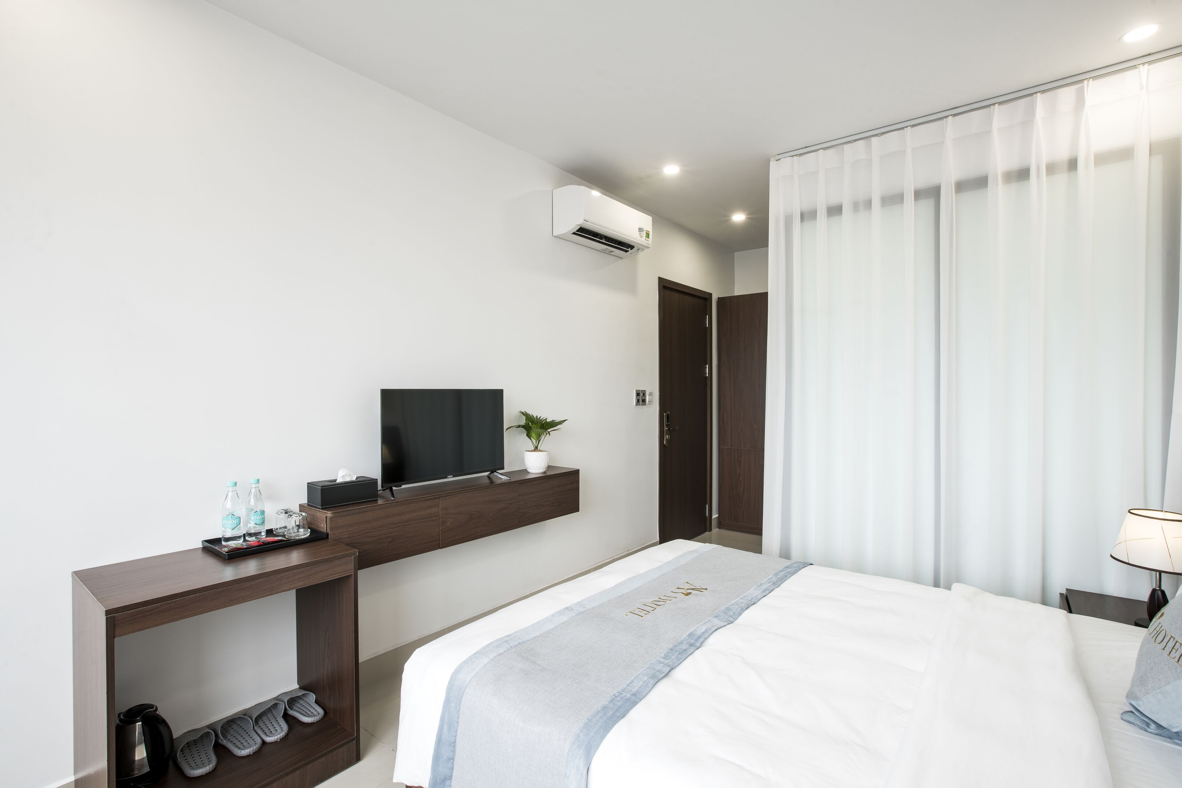 A modern hotel room with a bed, a TV on a wooden console, a shoe rack, and a window with curtains.