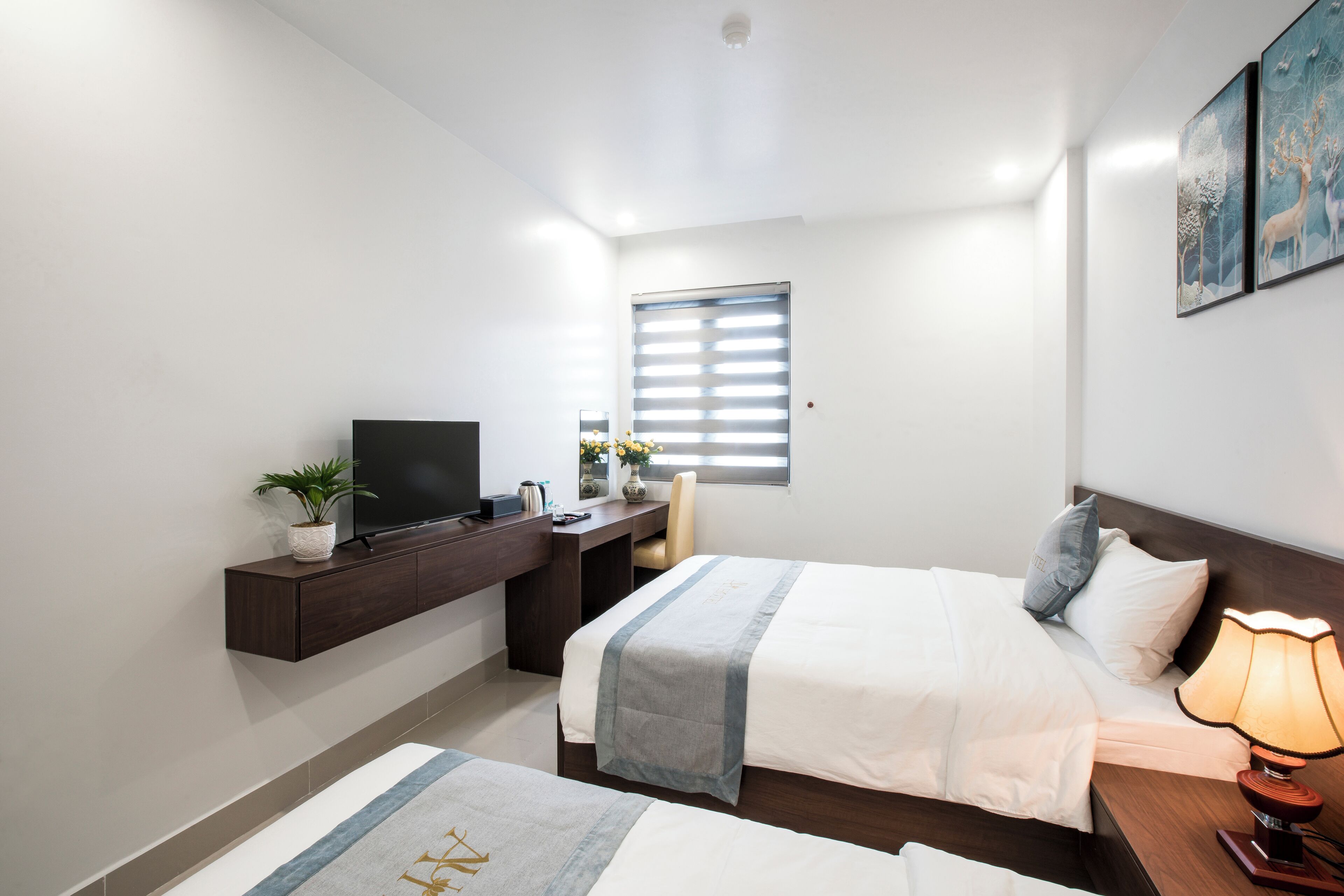 Una habitación de hotel con cama, mesita de noche con lámpara, escritorio con televisor, ventana con persianas y una obra de arte en la pared.