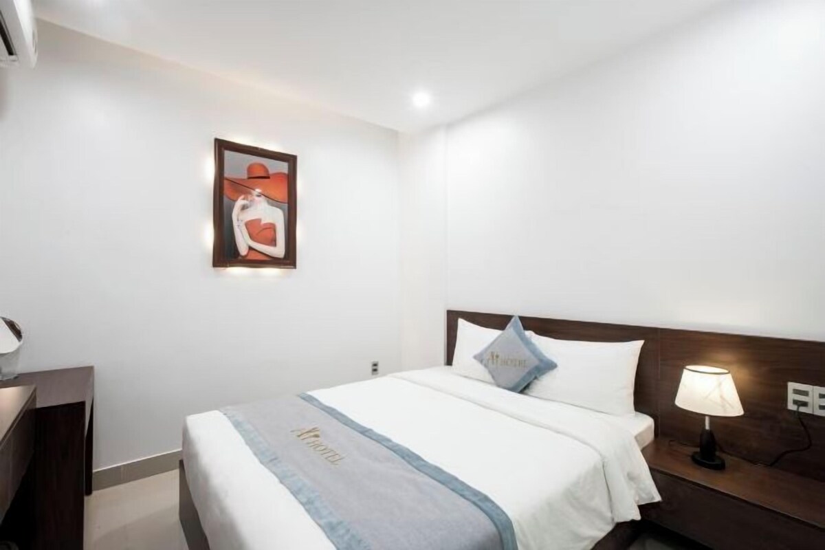 A hotel room with a bed, a nightstand with a lamp, a wall-mounted air conditioner, and a framed picture above the bed.